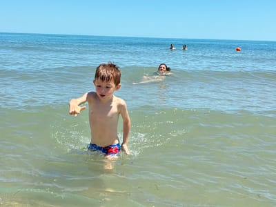 A boy is standing in the shallow water at the beach in Shkorpilovtsi, Bulgaria, looking towards the camera. He has brown hair and is wearing blue and red swim trunks. In the background, a woman is swimming in the ocean, looking towards the boy. Further in the background, two more people are swimming in the distance, and an orange buoy is visible. The sky is clear and blue, and the water is calm and clear, with gentle waves. The sun is shining brightly, creating dappled light on the water.