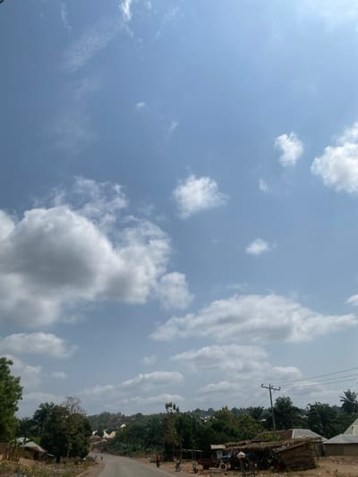 This image depicts a daytime scene along a paved road in Okura, Nigeria. The upper two-thirds of the frame consists of a light blue sky with numerous scattered white cumulus clouds. The road, appearing dry, runs through the lower third of the image, receding into the distance, flanked by dense green foliage, including various trees and visible palm trees. On the right side of the road, multiple rustic structures are present, including a roadside stall with a dark, possibly thatched, roof supported by visible wooden beams. Several individuals, indistinct due to distance, are observed near these structures, some standing beside what appear to be motorcycles or bicycles. Utility poles with overhead power lines parallel the road on the right. Additional buildings are partially visible amidst the vegetation in the mid-ground and on the left side of the road. The scene is illuminated by strong, even daylight.