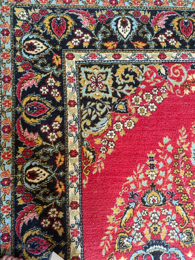 This is a close-up, overhead shot of a decorative rug with a red base color and intricate patterns. The rug features a border design with alternating sections of black and lighter colors, possibly cream or light blue, adorned with floral motifs. The central part of the rug displays a repeating pattern of stylized flowers and paisley-like shapes in various colors including red, blue, yellow, and white. The texture of the rug appears to be soft, possibly wool or a similar material, with a slight sheen from the fibers. There are no people or specific objects visible, and the scene is a static close-up of the textile. The lighting suggests it is daytime, but no specific time or weather conditions can be inferred. No text is visible in the image. The provided location context of Rybnitsa, Moldova, does not offer any specific visual cues within the image itself.