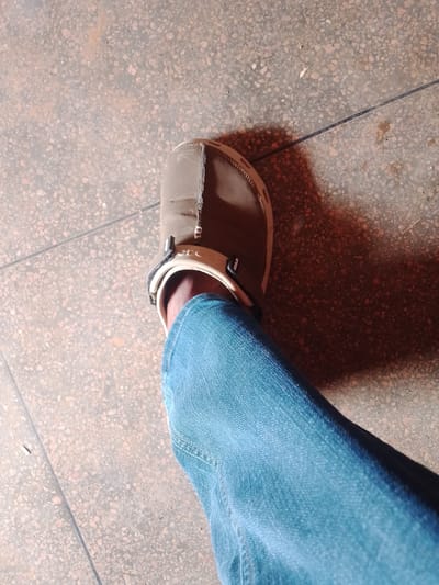 A close-up shot focuses on a person's right foot clad in a brown Croc-style shoe, paired with a blue denim jean leg. The foot is situated on a tiled floor. The location, Jama A, Nigeria, is not discernible from the image.