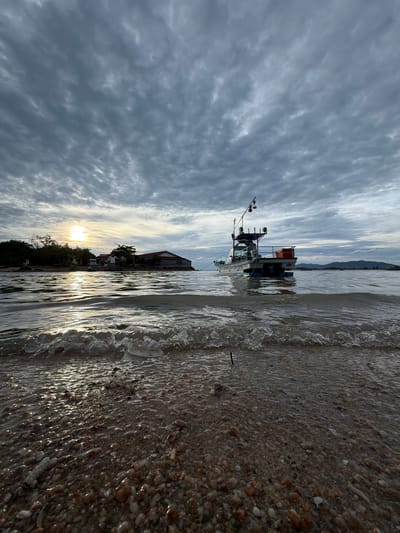 This is a vertically oriented, wide-angle photograph taken from a low perspective at the edge of a beach. The image is dominated by the sea on the right, with waves gently lapping at a shore covered in pebbles and small shells. The water is partially covering the shore, reflecting the sky and sunlight.

On the left side of the frame, the sky is filled with dramatic, textured clouds, with streaks of white and gray suggesting movement. The sun is visible low in the sky, casting a bright, diffused light that creates a shimmering effect on the water. A small boat with a cabin and a mast is anchored in the water. In the background, silhouetted against the sky and sun, is a tree-lined shore with a building.

The time of day appears to be late afternoon or early evening, given the low position of the sun. The weather is cloudy but bright. The scene suggests a tranquil coastal setting. No people are visible in the image. There is no visible text. The location cues suggest a coastal area in Ko Samui District, Thailand.