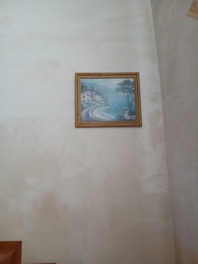 The image shows a framed painting on a wall. The painting depicts a coastal village scene with buildings and water. The wall is painted a light, neutral color and appears textured. The ceiling is visible at the top of the frame and has a similar textured finish. The overall lighting suggests an indoor setting. There is no visible text in the image. The composition focuses on the painting and its immediate surroundings on the wall.