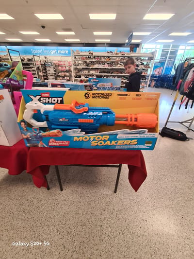 This is a medium shot, taken from a slightly elevated angle, of a toy water gun display in a retail store. The primary object in focus is a large, blue and orange water gun in its packaging, which is a bright yellow box. The text on the box reads "XSHOT MOTORIZED SOAKERS" and "GATLING PULSE". The toy gun is positioned on a table covered with a red cloth, suggesting it's part of a display. To the left of the water gun, a young boy with short brown hair is looking towards the toy. He is wearing a black hoodie and appears to be browsing.

In the background, shelves filled with various shoes are visible, indicating the setting is a shoe store or a department store with a shoe section. Above the shelves, a large blue sign with white text reads "Spend less get more," a common retail slogan. The store appears to be well-lit with fluorescent ceiling lights. The floor is a speckled gray material, typical of commercial spaces. The overall scene suggests a shopping environment, possibly during daylight hours given the lighting. The boy's expression is neutral, and there are no other people prominently featured in the image. The photograph also includes a watermark at the bottom right corner: "Galaxy S20+ 5G".