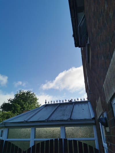 The image captures a view from ground level looking upwards towards a blue sky with scattered white clouds. On the right, a brick building with a dark roofline and an open window is visible. In the center, the roof of a conservatory with decorative finials is prominent. A large green tree can be seen behind the conservatory. The scene appears to be in a residential area in Ripon, United Kingdom, as indicated by the architectural style and the presence of mature trees. No people are visible in the image, and no actions or interactions are depicted. The environment suggests an outdoor setting on a clear day.