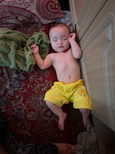 A baby is sleeping on a bed. The baby is wearing yellow shorts with a smiley face logo. The bed has a red patterned comforter and a green blanket. The baby is lying next to a beige piece of furniture, possibly a dresser or a bed frame. The scene appears to be indoors, in a bedroom. The location is in Kyzylorda, Kazakhstan.