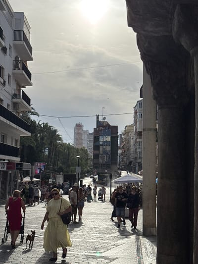 The image depicts a bustling street scene in what appears to be a Mediterranean city, likely Benidorm, Spain. The perspective is from under an overhang or archway, looking out onto a pedestrianized street. The sun is bright, suggesting it's daytime, and the sky is partly cloudy.

The street is paved with a patterned tile mosaic, typical of many Spanish towns. A diverse group of people are walking along the street, some in small groups, others singly. Many are dressed in summer clothing, indicative of warm weather. On the right side of the street, a woman in a light yellow dress, wearing a hat, is walking a small dog on a leash. Further ahead, another woman in a red dress is seen. The people's expressions are not clearly discernible, but the general atmosphere appears to be one of casual strolls and perhaps shopping, given the presence of shops and awnings visible in the background.

The background features multi-story buildings with balconies, characteristic of coastal resort towns. Some buildings are modern, while others have a more traditional facade. Palm trees are interspersed among the buildings, further reinforcing the warm climate location. Overhead, electrical wires are strung across the sky.

There is some visible text in the image, including what appears to be pricing information like "10€" on a sign, and potentially shop names or advertisements, though they are mostly illegible due to the image quality and distance. The overall scene conveys a lively, sun-drenched urban environment during the day.