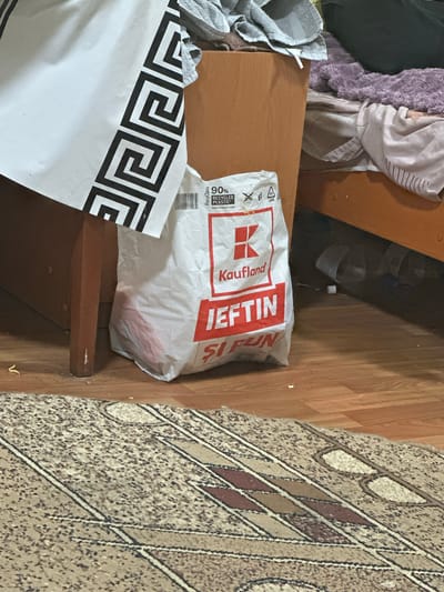 The image depicts an indoor scene, likely a bedroom or living area, with a focus on a white plastic shopping bag from "Kaufland" placed on a wooden floor. The bag is prominently displayed, with text in red lettering that appears to say "IEFTIN" (meaning "cheap" in Romanian) and "SI FUN". Next to the bag, a wooden table leg is visible, and to the right, a bed with bedding is partially seen. A portion of a patterned rug covers the floor in the foreground. A white sheet with a black Greek key pattern hangs on the left side of the image. The scene appears to be in a state of disarray, with various items scattered around. The overall impression is that of a casual, lived-in domestic space, possibly in a residential apartment or house within the city of Corlăteni, Moldova. No individuals are present in the image.