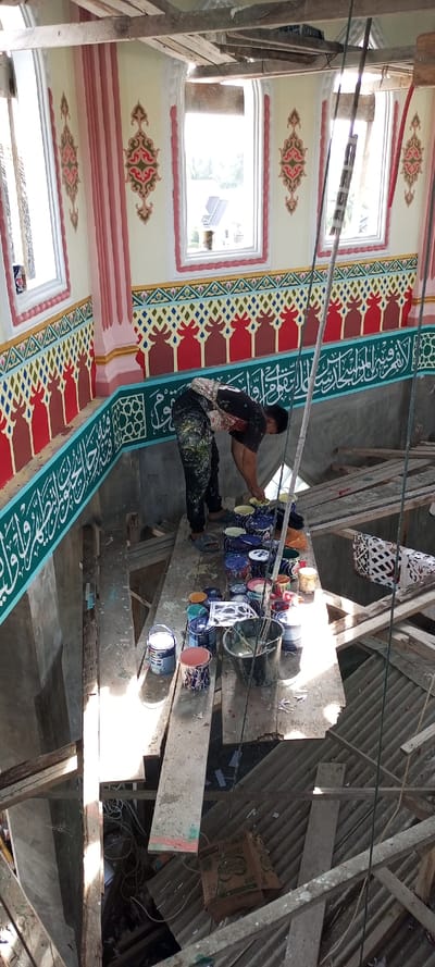The image depicts an interior scene during construction or renovation in the city of Kumbang, Indonesia. A male individual is positioned on a makeshift wooden scaffold, actively engaged in painting or paint preparation.

The individual, wearing a dark, paint-splattered t-shirt and similarly stained dark trousers and blue flip-flops, is bent over a collection of paint containers. A light-colored cloth or bandana is visible on his head. On the wooden plank scaffold around him are numerous paint cans and buckets of various sizes and colors, including several with blue and white patterns, a large white can labeled "Dulux," and a dark bucket containing brushes or stirring tools. Several stencils or templates are also visible on the scaffold.

Above the scaffold, the building's walls are extensively decorated. The upper section features cream-colored walls with four arched windows framed in reddish-pink. Through the windows, distant trees and a building roof are visible under bright daylight. Between and below the windows are intricate red, green, and cream ornamental patterns. Below these, a horizontal band exhibits a repeating geometric design in yellow, green, red, and blue. Further down, a section of the wall is painted solid red with scalloped motifs. A prominent turquoise-green horizontal band with white Arabic calligraphy runs across the wall directly above the scaffold level. Below this, the wall is a plain light grey, appearing unfinished. Exposed wooden beams and structural elements are visible near the ceiling.

Below the scaffold, a significant drop leads to a lower level of the construction site. This area contains additional wooden planks, corrugated metal sheets, a brown cardboard box, and loose ropes or cables. The scene is brightly illuminated by natural light entering through the windows, casting shadows on the scaffold and lower levels.