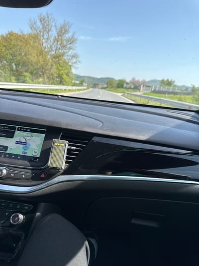 The image shows the interior of a car, likely in Daskotna, Bulgaria. The dashboard features a navigation screen displaying a route on ул. Иван Вазов, with a current speed of 76 km/h and a time of 10:29. An air freshener is attached to the air vent. The surrounding environment visible through the windshield includes a road, trees, and clear sky. The image appears to have been taken during a car journey.