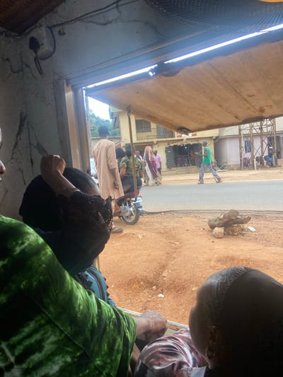 Captured from an interior perspective, the image presents a view of a street scene in Okengwe-Obehira, Nigeria. Several individuals are visible along the street, and at least one motorcycle is present. The roadside exhibits dirt and some wood debris. Inside the structure from which the image is taken, multiple people are partially visible. The interior structure appears weathered.