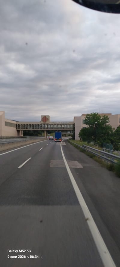 The image shows a highway scene in La Jonquera, Spain, with a large, modern building complex spanning over the road. A blue truck is driving on the highway, with other vehicles in the distance. The sky is overcast with clouds. The text at the bottom of the image indicates that the photo was taken with a Galaxy M52 5G on June 9, 2026, at 06:34. The building complex appears to be a service area or a commercial building, with the words "Porta Catalana" visible on its facade. The highway is a multi-lane road, with clear lane markings and guardrails on the side. Trees and vegetation are visible beyond the highway. The overall impression is that of a typical highway scene during daytime, with a significant architectural structure overhead.