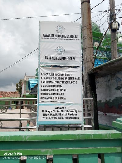 A white sign with blue and red lettering hangs on a metal fence post in front of a green building in Kecamatan Haurgeulis, Indonesia. The sign reads, “Yayasan Nur Ummul Qolbi” at the top and “TK IT Nur Ummul Qolbi” below. There are six bullet points on the sign, outlining the school’s curriculum, which includes “Baca Tulis Al-Quran (BTQ),” “Praktek Sholat Dhuha Setiap Hari,” “Menghafal Surat Pendek Juz 30,” “Manasik Haji Cilik,” “Bahasa Inggris Dasar,” “Bahasa Arab Dasar,” and “Pramuka & Olahraga.” The sign concludes with the school’s address: “Alamat: Jl. Raya Cinini Sumbermulya Blok Masjid Baitul Firdaus Rt 13 Rw 07 Kec. Haurgeulis”. The green building behind the sign appears to be a mosque or place of worship. A portion of the sign is obscured by a metal fence that runs across the front of the building. The sign is being photographed with a mobile phone held in portrait mode. The date and time, “23 Mar 2023, 12:43,” appears in the lower right corner of the image. The image is taken from a low angle, pointing upwards towards the sign.