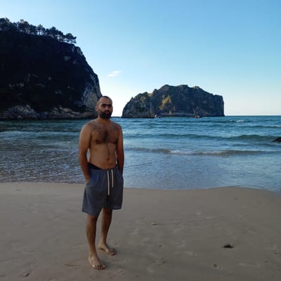 A man stands on a sandy beach in Ribadedeva, Spain, with the ocean and rocky cliffs in the background. The man is shirtless and wearing grey shorts. He has dark hair and a beard. The ocean has small waves, and the sky is clear and blue. There are trees on top of the cliffs. The overall scene is natural and serene.