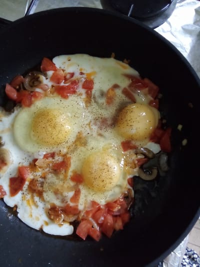A close-up overhead shot shows four eggs being fried in a black non-stick pan. The eggs are in the center of the pan, with their yolks mostly intact and the whites spread out. Diced tomatoes and sliced mushrooms are scattered around the eggs, and the entire dish is seasoned with black pepper. The pan is on a surface that appears to be covered with aluminum foil, possibly a stovetop. The scene is a domestic setting, likely a kitchen in a dacha settlement.
