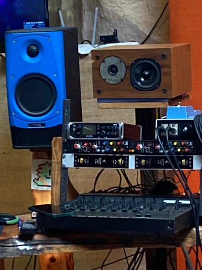 A brown wooden speaker sits above a blue and black speaker with a visible "PMC" logo. To the right, a rack-mounted audio interface with various inputs and outputs is stacked with other equipment, including a tall black mixer featuring numerous knobs and faders. Cables snake around the devices. A small pixel art sticker of a green dinosaur is visible underneath.