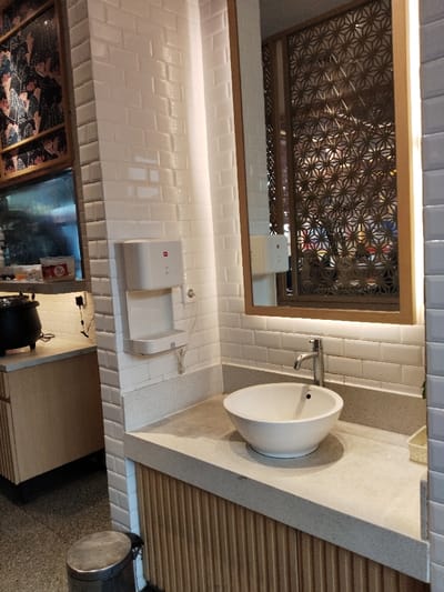 The image shows a bathroom sink in a restaurant located at the coordinates -6.301 latitude and 107.278 longitude in the city of Telukjambe, Indonesia. The sink is white porcelain and is mounted on a countertop made of light gray speckled material. The countertop is attached to a cabinet with wooden slats. Above the sink is a mirror reflecting a decorative wooden screen. The wall is tiled in white, and there is a white electric hand dryer mounted to the wall next to the sink. There is a metal garbage can in the foreground.  The space is clean and well lit.