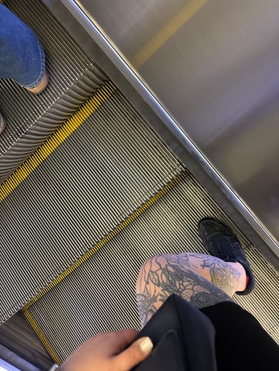 A high-angle shot shows the perspective of someone riding an escalator. The escalator has a silver handrail on the left side and a grooved metallic surface with yellow demarcation lines.
Visible on the right side is a leg with extensive tattoos, wearing a black shoe. A black bag is being held. On the lower left, a person wears blue jeans.
There is no specific information to link this scene directly to Newcastle upon Tyne, UK.