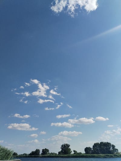The image depicts a serene natural landscape under a vast, clear blue sky dotted with scattered white cumulus clouds. In the foreground, a calm body of dark blue water stretches horizontally across the frame. Along the far bank of the water, a dense line of lush green trees and vegetation forms a natural horizon. The trees are varied in size and shape, with some appearing as large, mature specimens and others as smaller bushes. The overall impression is one of a tranquil day in a rural or park-like setting. The location is identified as Bilousivka, Ukraine.