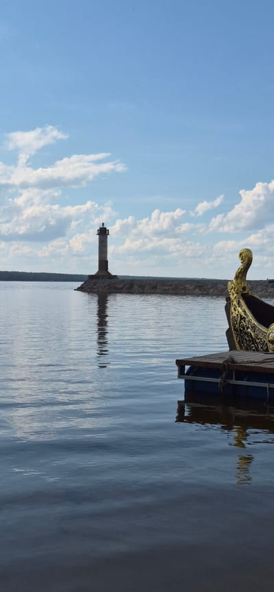In the city of Motornoe, Russia, a lighthouse stands on a stone pier against a sky filled with scattered white clouds and blue hues. The water is calm and reflects the lighthouse. In the foreground, the ornate golden prow of a boat is visible, tethered to a wooden dock. The scene captures a tranquil waterscape with a historical maritime element.