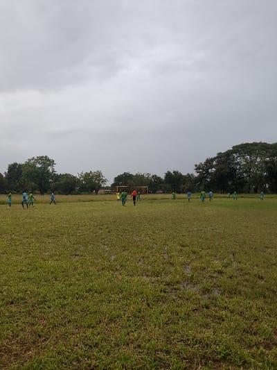 The image shows a grassy field with a group of people playing soccer. The scene takes place in Morogoro, Tanzania, at the coordinates -6.799 latitude and 37.63 longitude. The field is wet and has puddles of water. The sky is overcast with a grey cloud cover. In the distance, there are trees lining the edge of the field. The players are wearing blue and green uniforms. One person is wearing a red shirt. The field is surrounded by trees. The players are running and kicking a soccer ball. The players are interacting with each other. The people are engaged in a game of soccer.