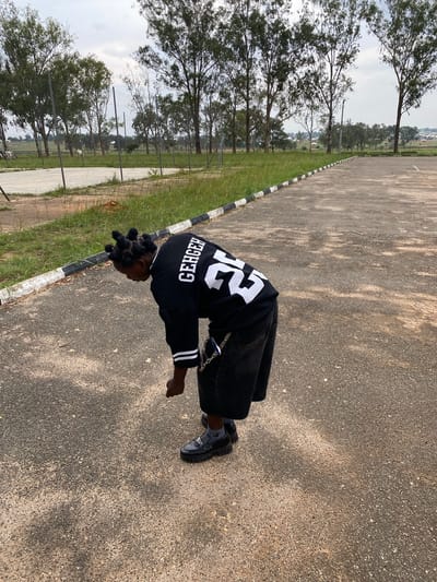 In Zawan, Nigeria, a person with braided hair is bending over on a paved surface. They are wearing a black jersey with white lettering and stripes on the sleeves, black shorts, and black loafers with silver accents. A silver chain is attached to their shorts. The background features a chain-link fence, tall trees, and a cloudy sky.