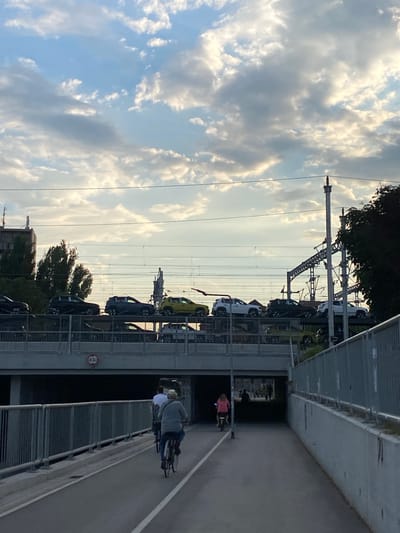 This is an eye-level, medium shot of a pedestrian and bicycle underpass in Subotica, Serbia, during the daytime. The sky is partly cloudy with sunlight breaking through.
In the foreground, a paved bicycle path runs through the underpass. Two cyclists are visible, heading away from the camera. The cyclist on the right wears a light-colored hat and a grey jacket, while the cyclist on the left wears a white shirt. Both are riding bicycles.
The underpass has concrete walls and a metal railing on the right side. The tunnel opening is visible in the distance, with more figures and cyclists entering it. A sign with "4.2m" is attached to the bridge structure above the underpass.
Above the underpass, a multi-level parking structure is visible, filled with cars. These include dark-colored SUVs and a yellow car. Overhead, train tracks and power lines are supported by metal structures. Buildings and trees are visible in the background on the left, and trees on the right. The overall scene depicts an urban environment with infrastructure for both vehicles and cyclists.