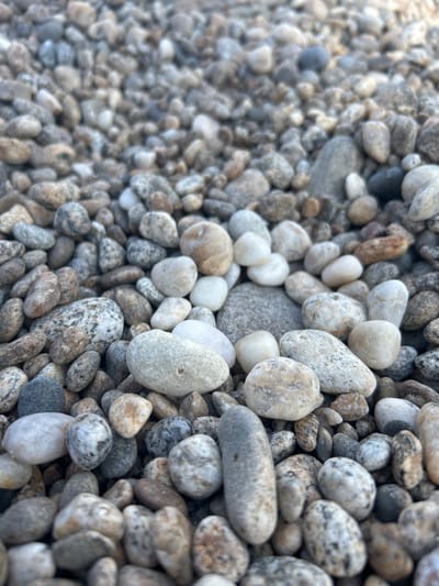 The image is a close-up, low-angle shot of a pile of small, rounded pebbles. The pebbles vary in color, ranging from white and light gray to darker grays, browns, and even some with reddish or tan hues. Many of the stones have a smooth, polished appearance, likely due to weathering or tumbling. The pebbles are densely packed together, creating a textured surface. The focus is sharp on the pebbles in the foreground, with the background becoming increasingly blurred, suggesting a shallow depth of field. The lighting appears to be natural daylight, casting subtle shadows that highlight the textures of the stones. There are no people or man-made objects visible in the frame, suggesting a natural setting such as a riverbed, beach, or path. The location in Tolbazikha, Russia, is not evident from the visual information within the image itself.