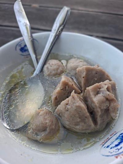 A close-up, top-down view shows a white bowl filled with broth and meatballs. Two silver spoons are resting in the bowl, with one spoon partially submerged in the broth. The broth is clear and slightly oily, with small bubbles on the surface. Several meatballs, some round and others cut into cubes, are visible in the broth. The bowl has blue and white insignia on the rim. The background appears to be a wooden surface, possibly a table. The image is shot outdoors, likely in a restaurant or food stall setting in Gianyar, Indonesia, indicated by the text "Cap Mobil" on the bowl rim.