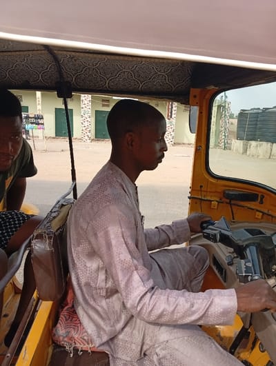 A man drives a yellow auto rickshaw, commonly known as a "keke," on a sunny day. He wears a light-colored, patterned shirt and matching pants. A passenger sits beside him, dressed in a dark shirt and patterned shorts. The rickshaw's interior features a patterned roof. The background shows buildings with green doors and water tanks, suggesting a semi-urban environment.