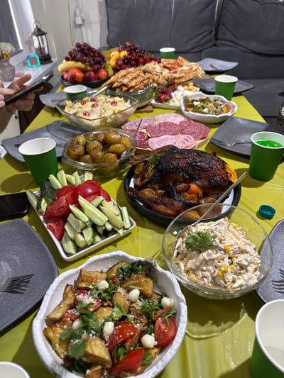The image shows a table laden with a variety of food items, suggesting a meal or gathering. The spread includes a roasted chicken, platters of sliced meats and cheeses, a salad, roasted potatoes, a fruit platter with grapes and peaches, and a dish of chopped tomatoes and cucumbers. Several green paper cups are visible, some filled with a green liquid. The table is covered with a lime green tablecloth, and part of a dark gray sofa is visible in the background, hinting at an indoor setting. A person's hands holding a smartphone are in the foreground. The lighting appears to be indoor lighting. The scene suggests a casual meal, possibly a celebration or family gathering. The location is identified as Sapronovo, Russia. No visible text is present.