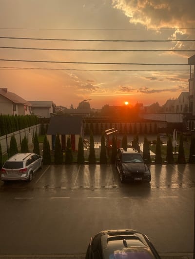 The image captures an outdoor scene during sunset in Novi Petrivtsi, Ukraine. The sun, a bright orange orb, is low on the horizon, casting a warm, golden light across the sky, illuminating scattered clouds. Power lines stretch horizontally across the upper portion of the frame, with buildings and trees visible in the background.
In the foreground, a paved area, possibly a parking lot or driveway, is wet, suggesting recent rain. Several cars are parked: a dark-colored sedan with license plate "MYD 781" is prominently featured, and a lighter-colored SUV with license plate "AK 6683 OX" is visible further down. The parked cars and the paved surface indicate a residential or commercial setting. The presence of manicured hedges and modern architecture suggests a well-maintained, possibly suburban or newly developed area. The overall mood is peaceful, with the soft light of the sunset creating a serene atmosphere. The time of day is clearly sunset, and the wet ground indicates recent precipitation.