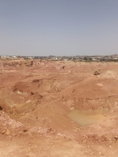 The image depicts an active construction site or quarry in Reifild-Ist, Nigeria. The terrain is characterized by uneven, reddish-brown earth with visible excavations and piled earth. Several shallow pools of murky water are scattered across the site. In the middle ground, a yellow excavator is visible, suggesting ongoing work. In the distance, a more developed area with residential buildings and some vegetation can be seen, indicating the proximity of a populated area. The sky is clear and light blue, suggesting a daytime scene. Several figures, appearing to be workers, are scattered across the site, some of whom are engaged in activities related to the excavation. A bright yellow hose or pipe is visible snaking across the ground in multiple sections of the image.