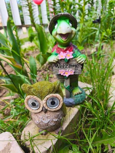 In a garden setting, two decorative figurines are positioned outdoors amongst green foliage and grass. In the foreground, a moss-covered owl figurine with large, reflective eyes sits on a light-colored stone. Behind and slightly to the right of the owl is a green turtle figurine wearing a hat and a red bandana. The turtle holds a wooden sign that reads "Welcome," adorned with pink flowers. The background shows a blurred white fence and more greenery. The scene appears to be in the city of Puchezh, Russia, given the context.