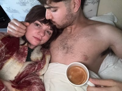 An indoor scene in Montepulciano, Italy, depicts two individuals in a bed. A man, positioned on the right, has dark hair, a beard, and a nose piercing. He is bare-chested, revealing chest hair and a thin gold chain around his neck. His head is tilted downwards, resting against the head of a woman on the left. The woman, with dark, shoulder-length hair and bangs, and a nose piercing, looks directly towards the viewer. She is covered by a red, white, and cream patterned blanket. The man's right arm extends around the woman. With his left hand, he holds a white mug containing a dark brown liquid with a layer of crema. Both individuals are situated on white bedding; a light-colored pillow with a blue and white patterned design is visible behind the woman.