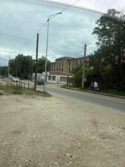 The image shows an urban scene in Silistra, Bulgaria. The foreground is a gravel roadside transitioning into a paved street. A building with light-colored walls and multiple windows stands alongside the road. Power lines and poles stretch across the sky. Several cars are parked along the street, partially obscured by trees. Two individuals, one wearing a white shirt, are walking along the edge of the road. The sky is overcast.