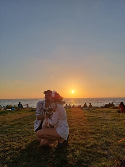 A couple is shown in the foreground, embracing and holding a small dog on a grassy area overlooking the sea at sunset in Palmachim, Israel. The man, wearing a patterned shirt and a cap, is partially obscured by the woman who is in a white lace dress and has flowers in her hair. The sun is setting over the ocean, casting a warm glow on the scene. In the background, other people are seated on the grass, enjoying the view. Beach chairs and bags are visible further down the coastline. The overall atmosphere is peaceful and romantic.