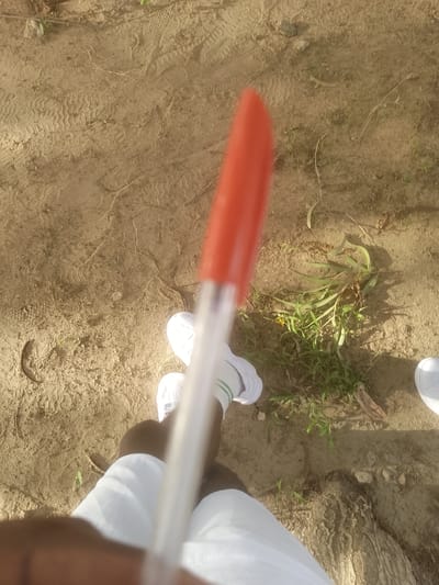 The image shows a close-up, overhead view of a person's legs and feet, wearing white shoes and white pants, standing on sandy ground. A blurry red and white object, possibly a pen, is held in the foreground, obscuring part of the scene. The ground is dry and dusty with visible tire tracks and some sparse green vegetation. The overall scene appears to be an outdoor, possibly rural, setting in Majemu, Nigeria.