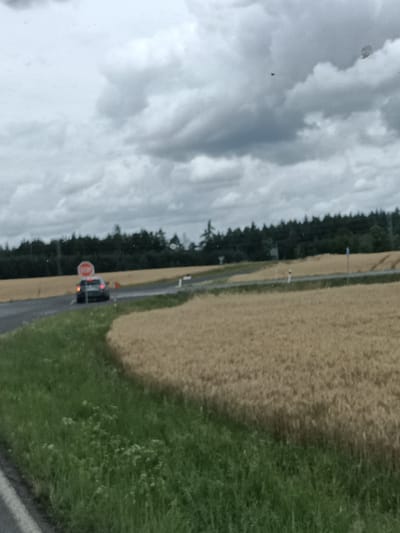 This is a medium-shot, landscape-oriented photograph taken from a vehicle, likely in motion. The foreground shows a grassy verge along the left side of a paved road. The road curves to the right, leading to a stop sign with a car stopped at it. Behind the road, a field of dry, golden crops, possibly wheat or barley, stretches towards a dense line of dark green trees that forms the horizon. The sky is overcast with heavy, grey clouds, suggesting a cloudy or potentially rainy day. There are two small, dark objects visible in the sky, one appearing to be a small, dark rectangle and another possibly a very distant, indistinct object. The lighting is diffused due to the cloud cover, and the overall mood is subdued. The visible text on the stop sign reads "STOP". The location is identified as Čistá, Czechia.