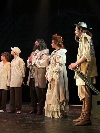 Five individuals, dressed in period costumes, stand on a stage with a dark, subtly patterned backdrop in Novoshakhtinsk, Russia. From left to right: a young person with dark curly hair wears a light tunic and dark trousers, with bare feet. Next, a person in a light-colored wide-brimmed hat, light tunic, dark trousers, and reddish-brown sandals stands, looking upwards. The central figure, a man with a dark beard and long hair, wears a ruffled light shirt, a dark patterned vest, a light brown tunic, dark trousers, and reddish-brown boots, with his hands clasped at waist level. Beside him, a woman with reddish-brown hair styled with feather-like adornments wears a layered light-colored dress with lace and ruffles, and a dark beaded necklace. On the far right, a man with long light-brown hair, a mustache, and a silver-colored wide-brimmed hat wears a light shirt, a light brown vest-coat, light brown trousers, and tall brown lace-up boots, holding a long, dark-bladed sword in a scabbard vertically in his right hand. All individuals are generally looking to the left or upwards, away from the viewer. The stage floor is dark and reflective.