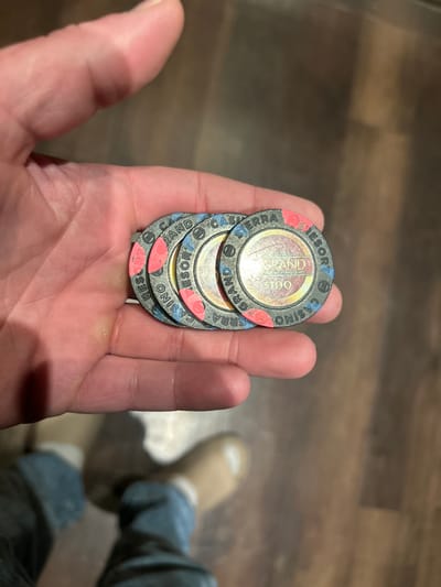 An adult human hand holds a stack of four casino chips against a dark brown, wood-patterned floor. The chips are uniformly designed, featuring a dark grey body with blue and red accents on the outer rim. Visible text on the chips clearly identifies them as belonging to the "GRAND SIERRA CASINO RESORT," located in Reno, Nevada, United States of America, and each chip displays a "$100" denomination. The chips exhibit signs of wear and surface dust. Below the hand, the lower legs of a person are partially visible, clad in blue denim jeans and light brown boots.