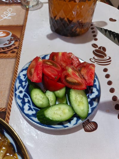 A close-up, eye-level shot shows a plate of sliced tomatoes and cucumbers, presented on a white tablecloth adorned with a repeating coffee cup and coffee bean pattern. The plate itself is blue and white with an ornate design. Behind the plate, a patterned glass pitcher filled with dark liquid, possibly tea, is visible. A serrated knife with a black handle lies to the right of the pitcher. To the left of the plate, a portion of a larger placemat with a coffee cup design is visible. The scene appears to be set on a table in a home or restaurant, likely in Urgench, Uzbekistan, given the traditional Uzbek patterns on the plate.