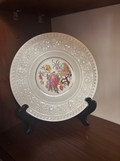 The image displays a cream-colored decorative plate, approximately 10-12 inches in diameter, positioned upright on a black, two-pronged plate stand. The plate features a central polychromatic floral design, including large pink blooms, smaller yellow and orange flowers, green leaves, and brown branch-like elements, enclosed by a thin green border. The wide rim of the plate is embossed with a repeating pattern of scrolled motifs and a continuous band of ovoid shapes. The plate and stand are situated on a dark brown wooden shelf or surface. The immediate background consists of dark brown wooden walls on the left and top, with a lighter-toned wall on the right. Two silver-colored L-brackets are visible in the upper left corner of the wooden enclosure. No individuals are present, and no active interactions are depicted. The specific location within the city of Monterey, United States of America, cannot be determined from the visual evidence provided.