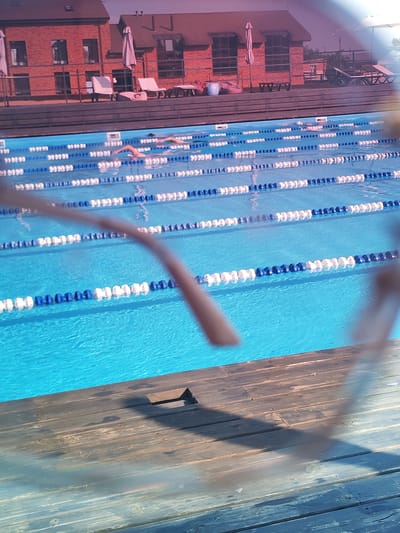 The image captures a scene at a swimming pool in Pirogovo, Russia. In the foreground, a wooden deck is visible with planks running horizontally. Beyond the deck, the bright blue water of a swimming pool fills the frame. Several swimming lanes are clearly marked with alternating white and blue buoys. In the middle distance, two swimmers are actively engaged in swimming strokes, with their bodies partially submerged in the water. In the background, a red brick building with dark windows and tiled roofs stands, accompanied by outdoor furniture, including lounge chairs and umbrellas, suggesting a recreational area. The lighting suggests it is daytime.