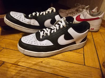 This image shows a pair of black and white Nike sneakers in the foreground, positioned on a wooden floor. Behind them, another pair of white Nike sneakers with red accents is visible. The sneakers appear to be worn. The environment is indoors, likely a room with natural lighting, as suggested by the shadows on the wooden floor. There are no people in the image. The scene is a close-up shot of footwear, with no discernible landmarks or features that would indicate a specific location in Vilnius, Lithuania.