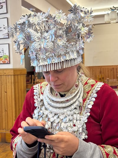 An adult female, light-skinned, is seated indoors in Fenghuang, China, wearing traditional ethnic attire. She is adorned with an elaborate, multi-tiered silver headpiece featuring floral, butterfly, and figurative motifs, along with several stacked silver neck rings and a broad silver chest ornament with dangling elements. Her main garment is a dark red, long-sleeved top extensively embroidered in gold and trimmed with silver disks. A grey undershirt is visible at the wrists. Her head is bowed as she interacts with a black-cased smartphone held in both hands. The background features light-colored wooden wall paneling and framed pictures displaying Chinese text and food visuals, indicative of a restaurant or cultural establishment.
