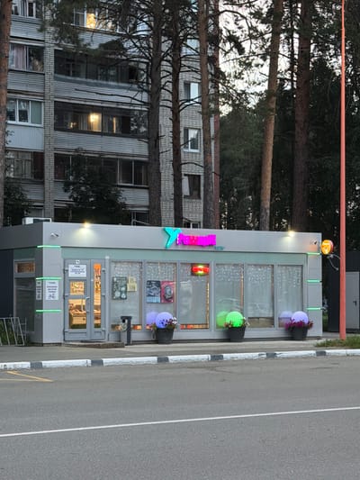 The image shows a small commercial building with a bright pink and blue sign that says "XAN". The building has large windows with decorative lights inside and two potted plants on the outside. There's an orange pipe running above the building. In the background, a tall apartment building is visible, with trees in front of it. The scene appears to be an outdoor setting, possibly a street or a parking lot, given the asphalt and road markings. The lighting suggests it's late afternoon or early evening, with artificial lights from the building and the sky indicating dusk. The overall mood is somewhat quiet and perhaps a bit whimsical due to the colorful lighting. There are no people visible in the image. The visible text on the building includes the prominent "XAN" sign and some smaller signs on the doors, one of which appears to have Cyrillic text. The location is identified as Zarechny, Russia.