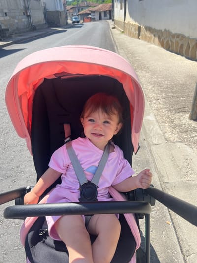 The image shows a baby sitting in a pink stroller on a street in Elena, Bulgaria. The baby is wearing a pink dress and is smiling at the camera. The stroller is black with a pink canopy. The street is paved and lined with buildings. A car is visible in the background. The sun is shining, casting shadows on the street.