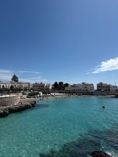 This image depicts a vibrant coastal bay, identifiable as Santa Maria al Bagno, a locality within the city of Nardò, Italy, observed on a clear, sunny day.

The foreground is dominated by calm, exceptionally clear turquoise-blue seawater, revealing the rocky seabed in shallower areas and reflecting sunlight with visible ripples. A dark rocky outcrop is present in the lower right corner, extending from the unseen immediate foreground. To the left, a natural rock formation and a constructed stone retaining wall delineate the water's edge, ascending to an elevated promenade.

The bay itself is populated by numerous individuals swimming, wading, and standing in the water. In the mid-ground, a person is visible seated on a red stand-up paddleboard.

Beyond the water, a crescent-shaped sandy beach stretches, densely occupied by sunbathers and beachgoers. A multitude of white and blue beach umbrellas are set up across the sand, providing shade. The beach transitions into a rocky shoreline on both the left and right sides of the bay.

Behind the beach and promenade, a series of multi-story buildings with various architectural styles and muted colors (white, cream, light pink, and ochre) line the coast, forming a small town or village. Green foliage from trees is visible interspersed among the structures and along the promenade. The sky above is a deep, unblemished blue with minimal white cumulus clouds visible only on the extreme right horizon. The scene is illuminated by strong, direct sunlight, indicating midday conditions.