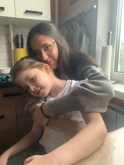 Two people, likely a mother and daughter, are captured in a warm embrace. The older person, wearing glasses and a grey cardigan over a white shirt, holds the younger person closely. The younger person, with light brown hair, looks towards the camera with a gentle smile, her arms wrapped around the older person's neck. The scene appears to be indoors, possibly a kitchen, with white cabinets and wooden countertops visible. A roll of paper towels hangs on a metal rod above a window, suggesting a domestic setting. The lighting suggests it could be daytime. The overall mood is affectionate and intimate, conveying a sense of familial love and comfort.