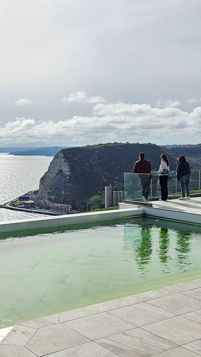 Three people stand on a building's edge overlooking a sea view in Kavarna, Bulgaria. A man in a burgundy shirt stands closest to the camera, then a woman in a white shirt, and finally another person with dark hair and a dark and light patterned shirt. They stand behind a glass barrier, with a decorative pool of water to the right and tiled decking. A large cliff with foliage and some buildings is centered behind them, with the sea visible on the left. The sky is partly cloudy.